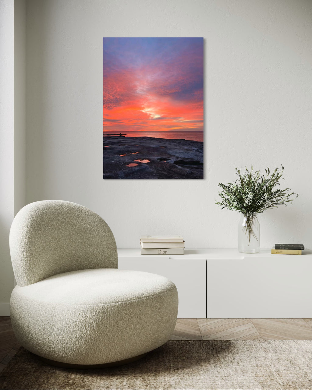 Modern chair in a room with a sunset painting on the wall and a vase with flowers on a cabinet.