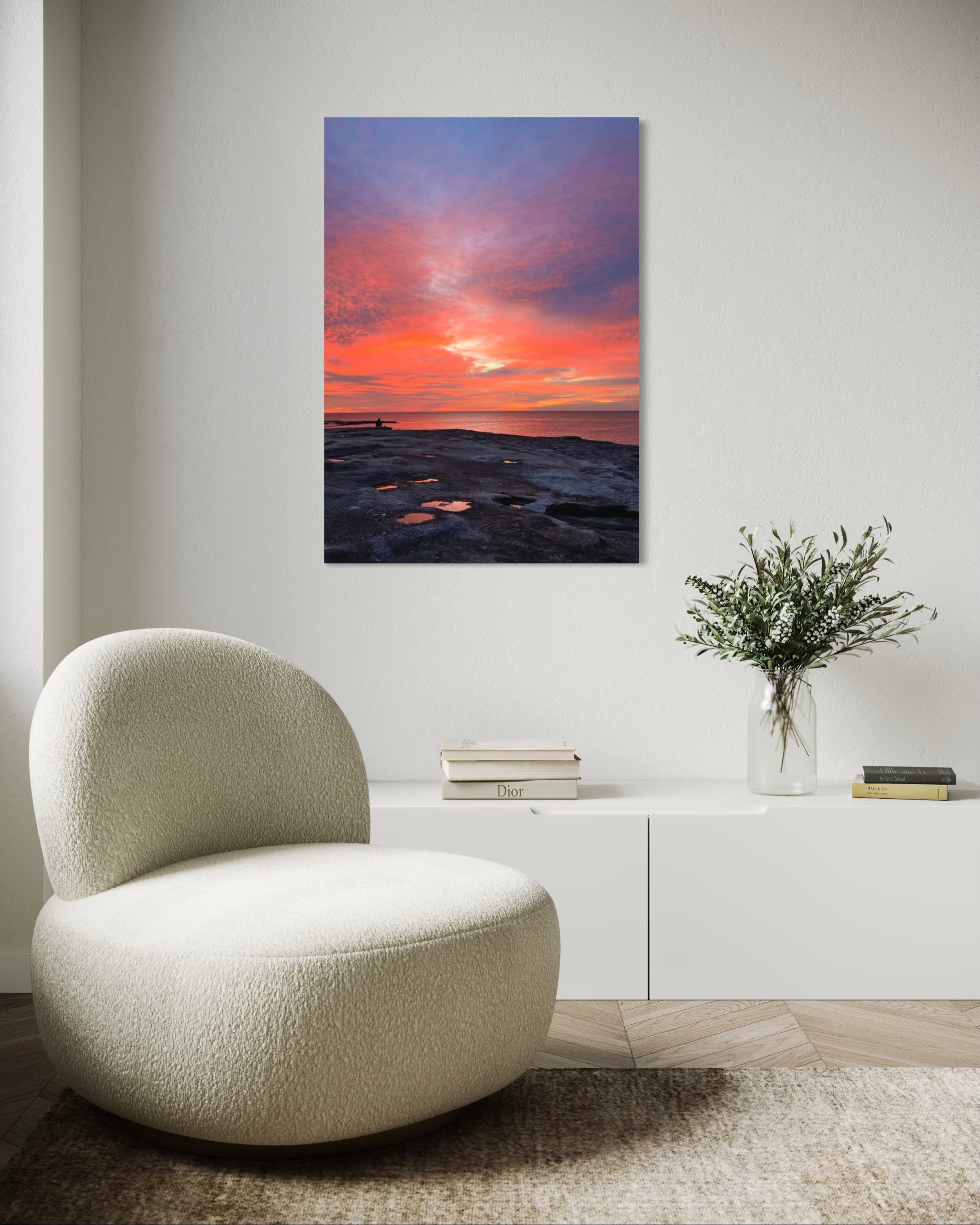 Modern chair in a room with a sunset painting on the wall and a vase with flowers on a cabinet.